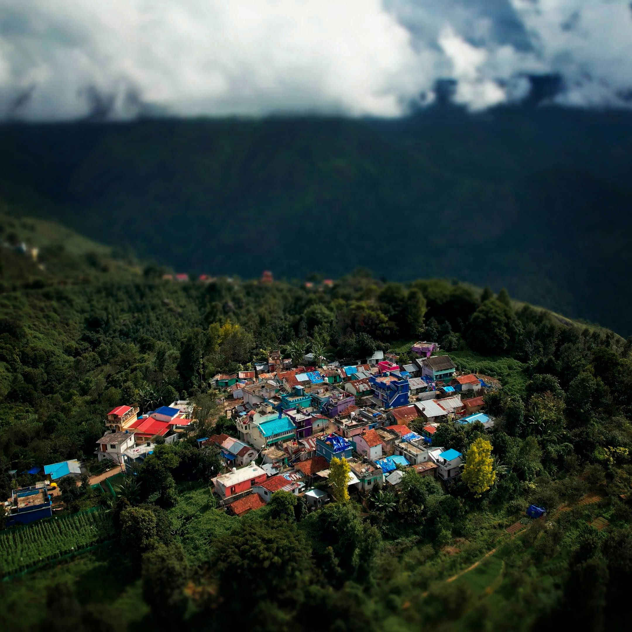 Drone shot of a vibrant village surrounded by dense greenery and mountains under a cloudy sky.